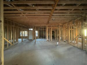 Unfinished interior of a building with exposed wooden framing and concrete floors. Ceiling beams are visible, and the room is well-lit by natural light from small windows.