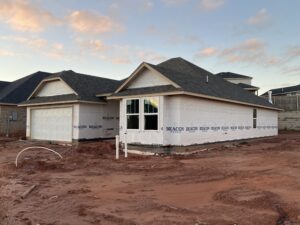 A partially constructed house with a garage, covered in weatherproof sheathing, surrounded by bare, red dirt. Cloudy sky in the background.