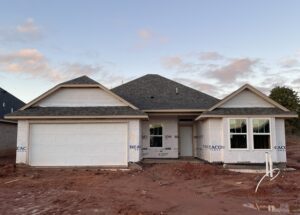 A newly constructed single-story house with a gray roof, partially covered in building wrap, and a double garage door. The ground is bare, with loose soil in front.