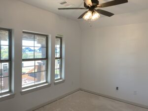 An unfurnished room with white walls, three windows, and a ceiling fan with lights. The floor appears bare and unfinished.