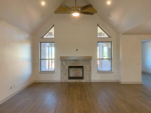 A spacious living room with wooden floors, a fireplace, and a ceiling fan. The room features two large windows and vaulted ceilings.