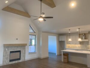 A modern, unfurnished living room with high ceilings, wooden beams, a ceiling fan, fireplace, large window, and an open kitchen with pendant lights and white cabinetry.