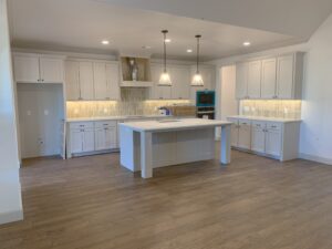 A modern kitchen with white cabinets, a central island, pendant lights, and wooden flooring.