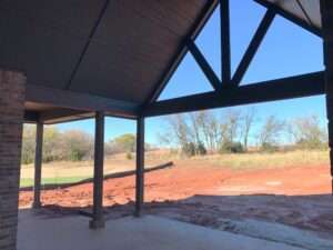 Covered patio with wooden beams overlooks a yard with red soil and patches of grass. Trees line the background under a clear blue sky.