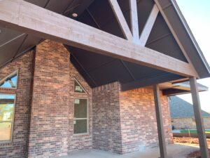 Brick house exterior with wood beams supporting a sloped black roof. Triangular window and another standard window visible. Construction materials on the ground.