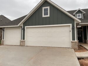 A green house with cream-colored trim has a three-car garage. The driveway is unfinished and the sky is overcast.