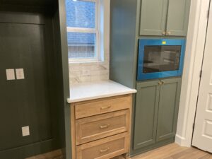 Kitchen corner with green cabinetry, a built-in microwave, and light wood drawers. White countertop and tile backsplash below a window.