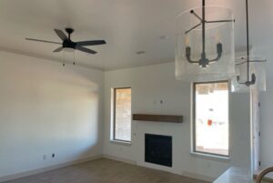 Empty living room with a ceiling fan, minimalist fireplace, and two pendant lights. Large windows provide natural light. Room has white walls and a wood-toned floor.