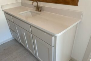 A white bathroom vanity with six drawers, a built-in sink, and a bronze faucet, set against a beige tiled floor and white walls.