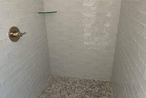 A tiled shower with light gray subway tiles on the walls and small pebble tiles on the floor, featuring a glass shelf and a metal shower control handle on the left wall.