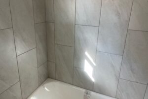 Bathroom corner with light gray tiled walls and a white bathtub. Sunlight shines through a window, creating a bright spot on the tiles.