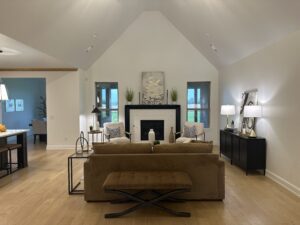 A modern living room with a high ceiling features a brown sofa, a bench, a fireplace, two windows, and a side table with lamps. The space opens into a kitchen area.