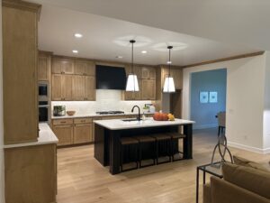 A modern kitchen with wooden cabinets, a black island with stools, pendant lights, and stainless steel appliances. A white backsplash and decorative items are on the counters.