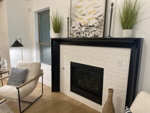 A modern living room with a black-framed fireplace, white brick surround, artwork, and plants on the mantel. A beige chair and floor lamp are nearby on a wooden floor.