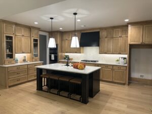 A modern kitchen with wooden cabinets, a central island with seating, pendant lights, and a white backsplash. Decorative pumpkins are placed on the island.