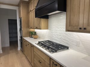 A modern kitchen with wooden cabinets, a black range hood, a gas stovetop, and white textured backsplash. Illuminated by under-cabinet lighting, a hallway and pantry are visible in the background.