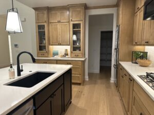 Modern kitchen with wooden cabinets, white countertops, a black sink, and a large island. Built-in appliances and shelving are visible in the background.