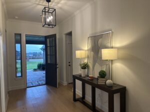 A hallway with hardwood floors, a console table with lamps and decor, and an open door showing a view of a porch and grassy yard.