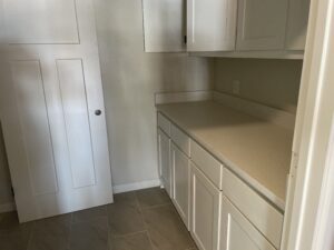 A white kitchen cabinet set with a white countertop and an adjacent door leaning against the wall. The floor is tiled in gray.