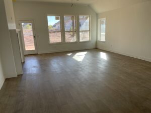 A spacious, empty room with large windows and wooden flooring, featuring white walls and a door leading outside. Natural light brightens the room.