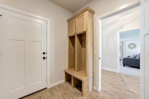 Entryway with a white door and a wooden storage bench with cubbies. The floor is light wood, and there's an open doorway leading to a living room with a gray sofa.