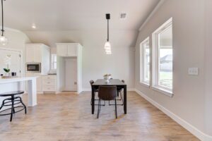 A bright, modern kitchen with light wood flooring, white cabinets, a black table with four chairs, and large windows.