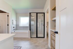 Modern bathroom with glass shower, white bathtub, and neutral tile flooring. A towel hangs by the tub, and built-in shelves are visible near a white door.