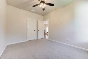 Empty room with beige walls, carpeted floor, and a ceiling fan. Two white doors with black knobs are on one wall, and a doorway opens to another room.