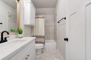 A modern bathroom with a white vanity, black faucet, a potted plant, a toilet, and a bathtub with a beige and gray shower curtain.