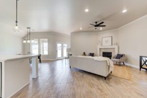 Spacious living area with light wood flooring, a modern kitchen, pendant lighting, a sofa, armchairs, and a fireplace. Large windows provide natural light.