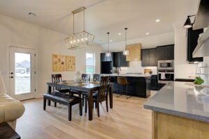 Modern kitchen and dining area with wooden floors, black cabinetry, and a large rectangular dining table with a bench and chairs. Pendant lights hang above the island. Natural light filters in.