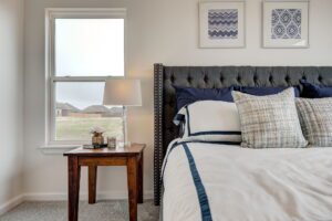 A neatly made bed with blue and white pillows is next to a wooden side table with a lamp and decor. Two framed artworks hang on the wall above the bed. A window is on the left.