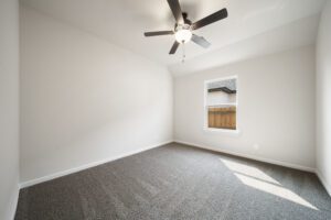 Empty modern room with gray carpet, white walls, a ceiling fan, and a window showing a wooden fence outside.