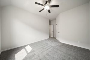 An empty, brightly lit room featuring gray carpet, white walls, and a black ceiling fan above, with two closed white doors on the opposite wall.