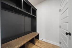 A modern mudroom with a wooden bench, black cabinets, hooks for coats, and a white door on the right.
