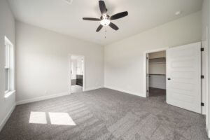 Empty room with gray carpet, white walls, ceiling fan, and open white door leading to another room. sunlight streams through window.