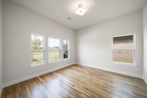Empty room with hardwood floors, white walls, and multiple windows allowing natural light, including one full-length and one smaller window.