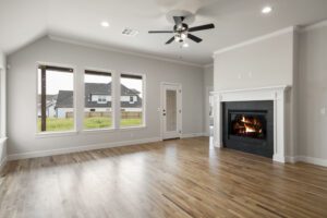 A spacious, modern living room with hardwood floors, a lit fireplace, and large windows overlooking a residential neighborhood.
