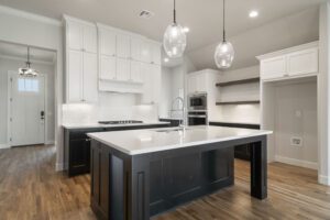 A modern kitchen with white cabinets, a dark island, stainless steel appliances, and pendant lights, featuring hardwood floors.