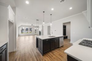 Spacious modern kitchen with a large island, pendant lights, and wooden floors, leading to a living area with ample natural light.