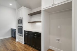Modern kitchen corner with black and white cabinetry, built-in appliances, floating shelves, and hardwood flooring.