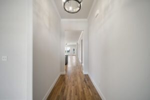 A modern home interior showcasing a long hallway with hardwood floors, white walls, and a simple overhead chandelier.
