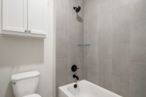 A modern bathroom with gray tiles, featuring a white toilet and a bathtub with black fixtures, and a built-in shelf on the wall.