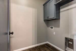 A small, modern utility room with gray cabinets, dark wood flooring, a thermostat on the wall, and a white door.