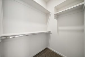 Empty walk-in closet with white shelves and hanging rods on either side, and beige carpet flooring.