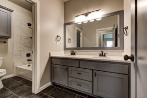 Modern bathroom featuring gray cabinets, double sinks, a large mirror, white marble walls, and a peek into the adjoining toilet area.