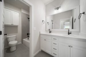 A modern bathroom featuring a double vanity with white cabinets, two sinks, a large mirror, black accessories, and a separate toilet area with gray tiles.