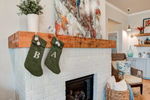 Two green christmas stockings marked "b" and "a" hanging on a rustic wooden mantelpiece above a white brick fireplace, with a vibrant painting and cozy seating area in the background.