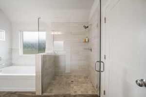 Modern bathroom with a glass-walled shower, bathtub, and frosted window. light gray tiles cover the walls and floor.