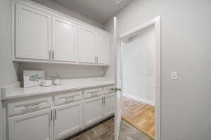 A modern laundry room with white cabinetry and countertops, featuring an open door leading to a room with hardwood flooring.